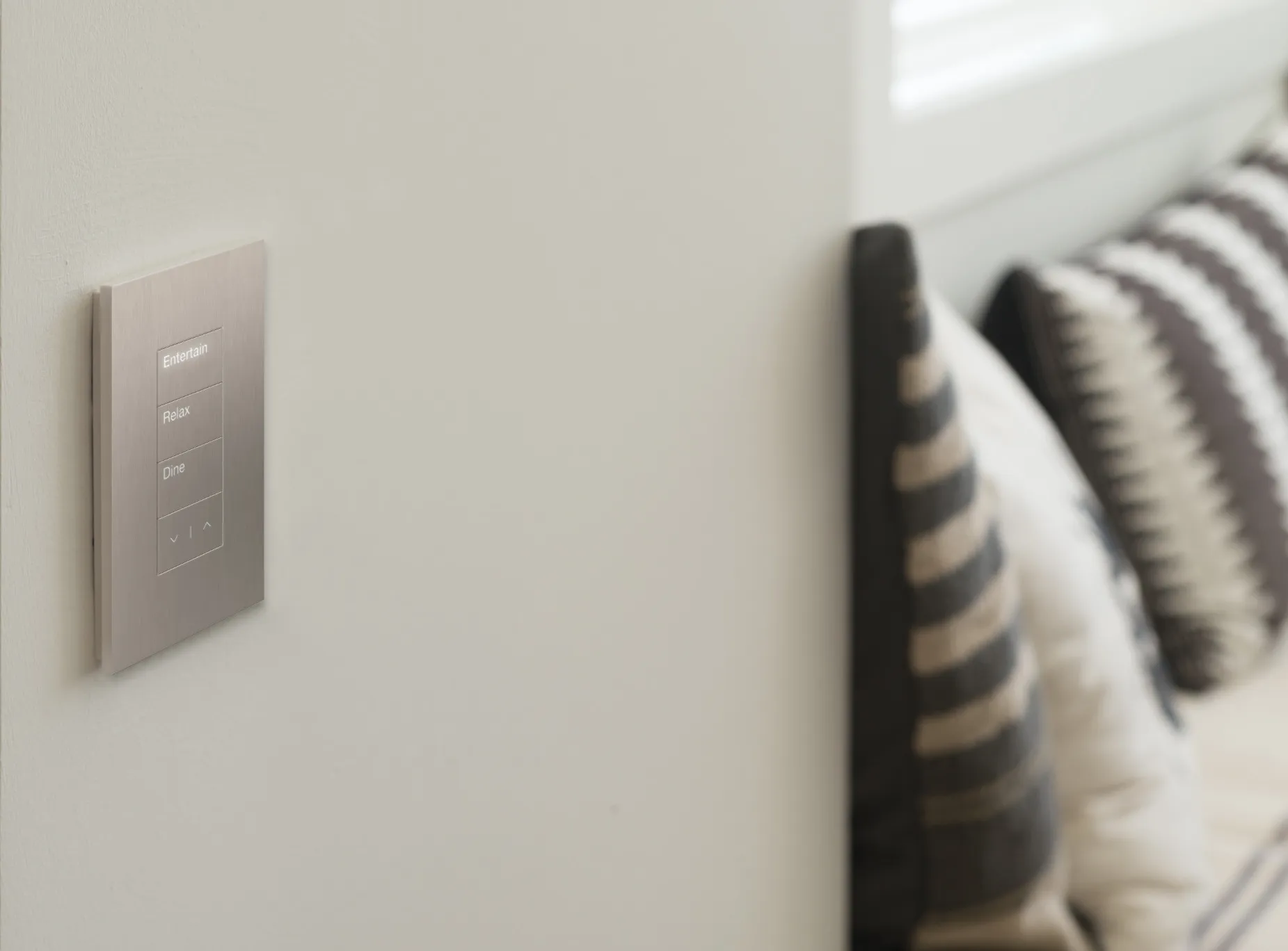 Lutron scene keypad on a neutral wall with brushed aluminum finish, showing Entertain, Relax, and Dine buttons with raise and lower controls — soft pillows visible in the background